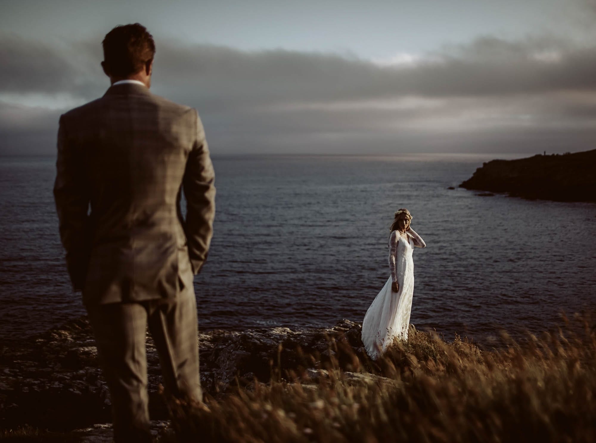 A groom in the foreground watches his bride standing on coastal rocks in the last light