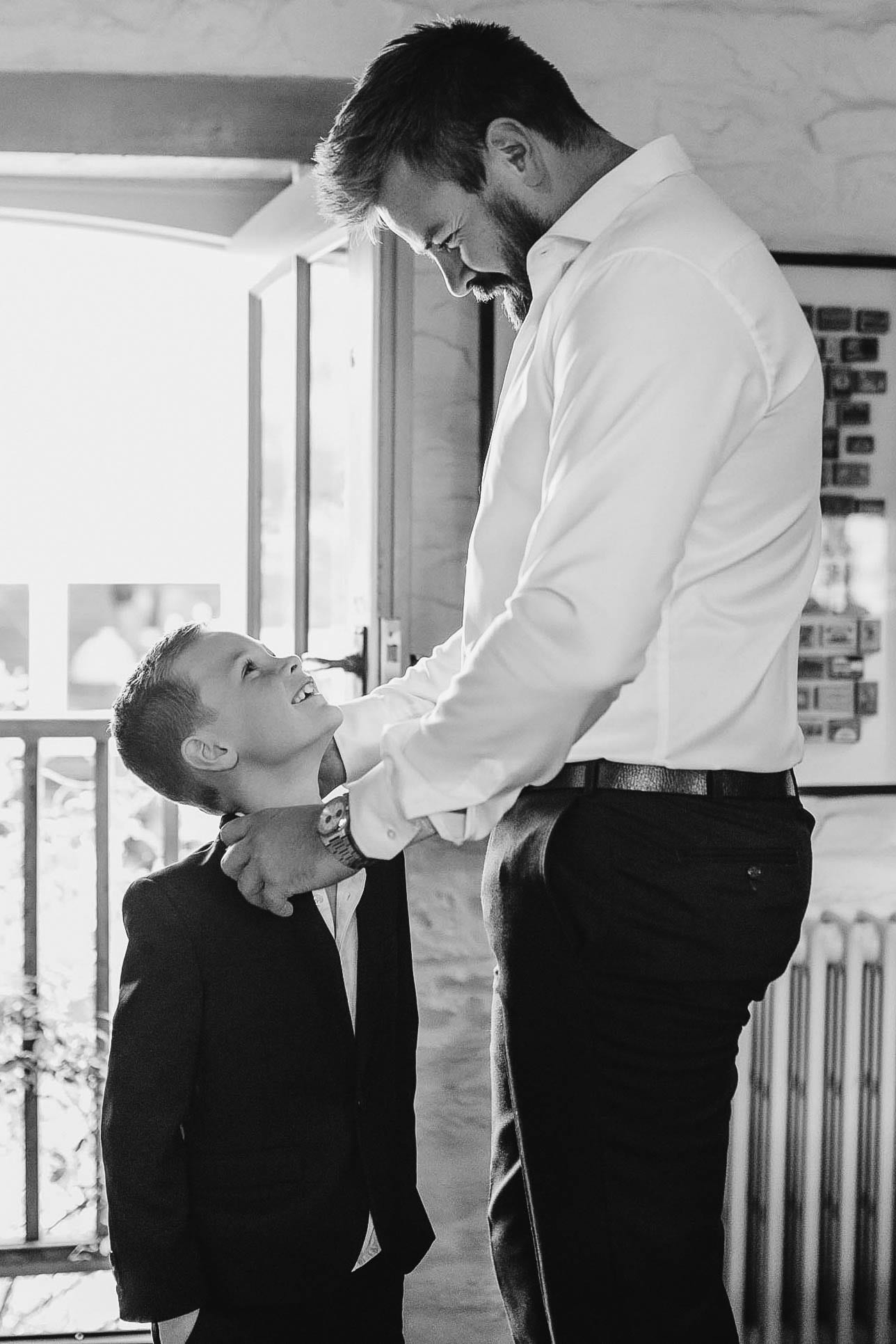 By an open doorway, a father straightens his young son's collar; the boy grins up at him