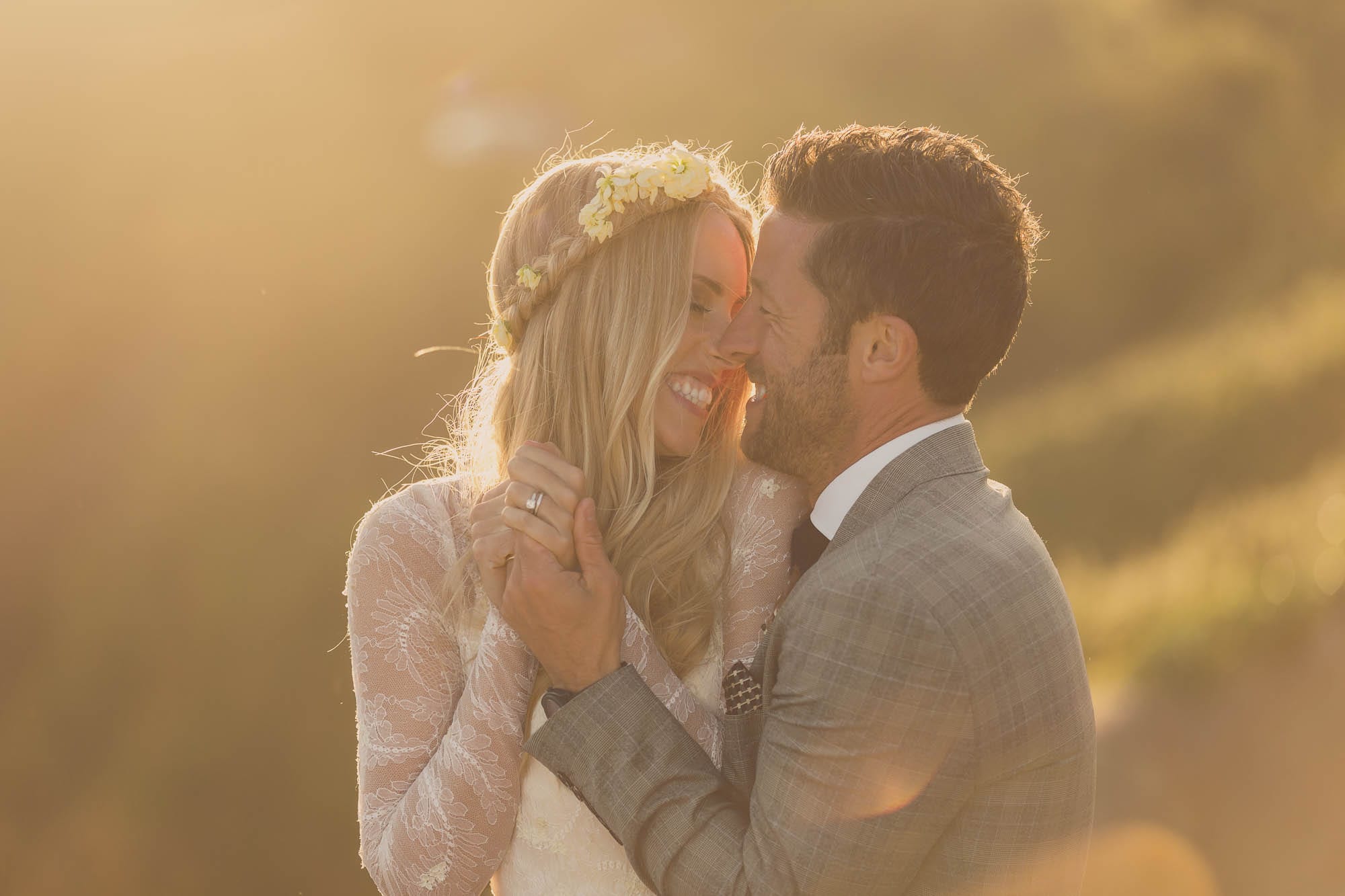 A couple laugh close together, backlit by golden evening sun, hands entwined