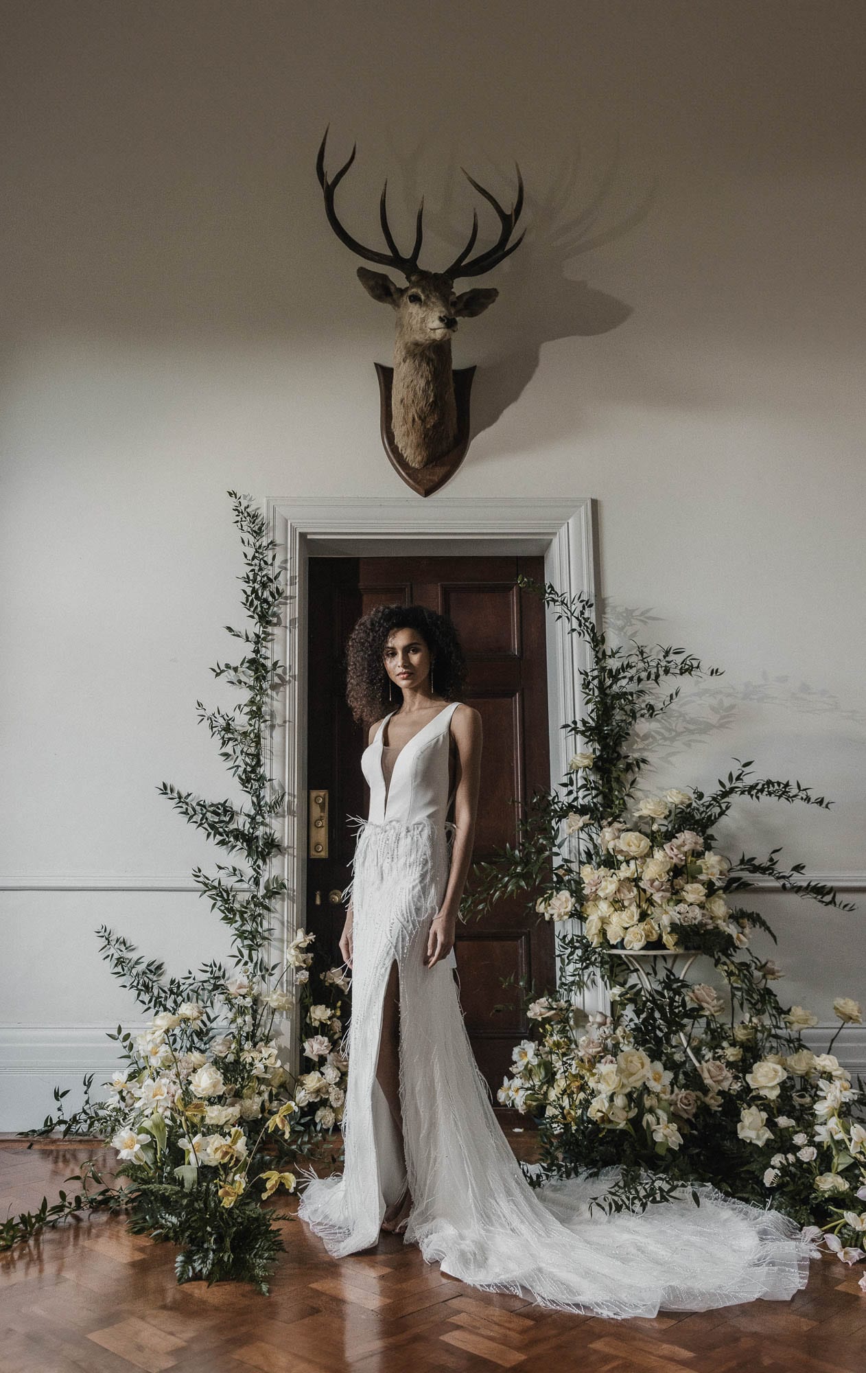 A bride stands framed by a doorway and flowers beneath a mounted stag's head